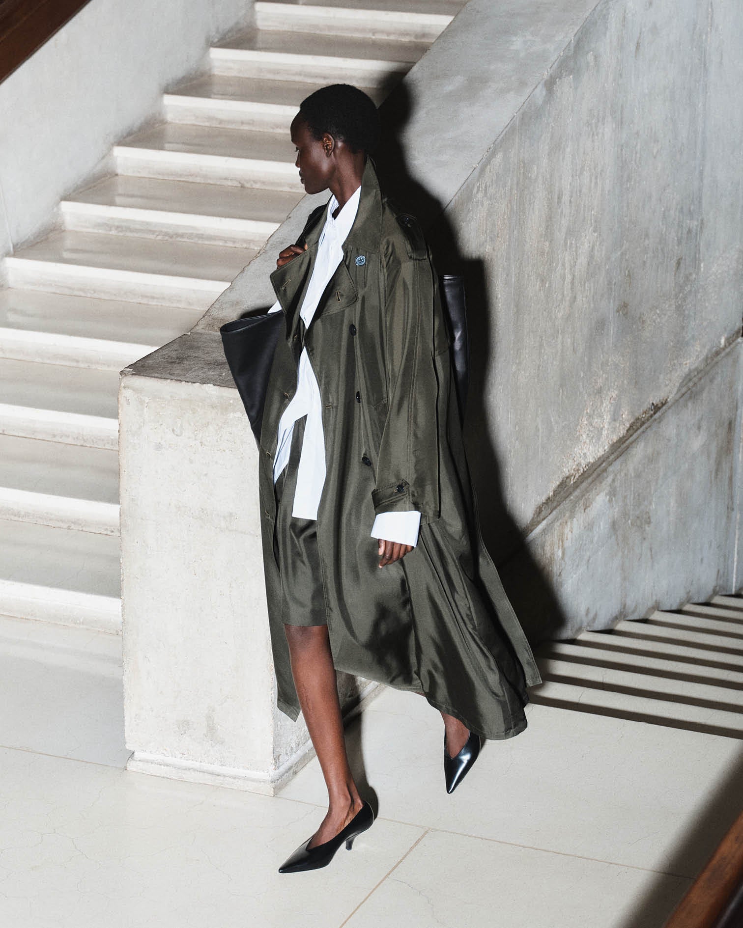 Model is photographed against brutalist architecture wearing a fluid green silk trench 