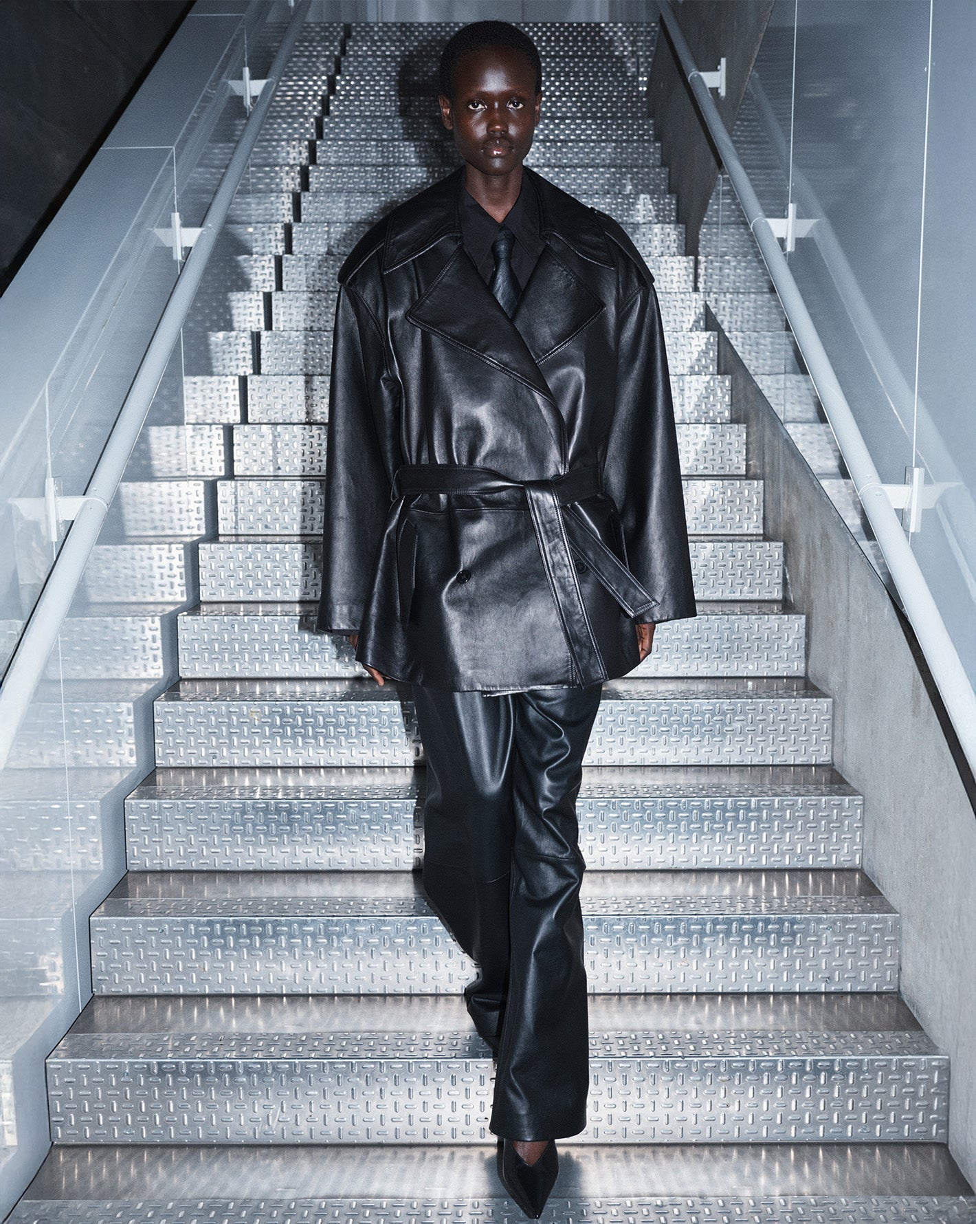 The Frankie Shop model walking down metal stairs wearing a leather trench and sleek leather pants. 