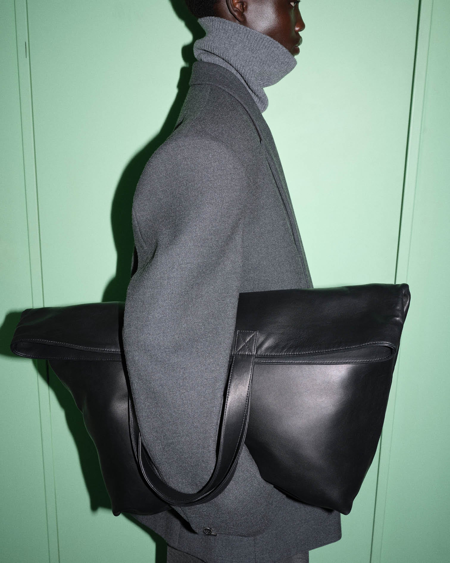 Model photographed against a green backdrop wearing a grey tailored suit holding a large black leather bag